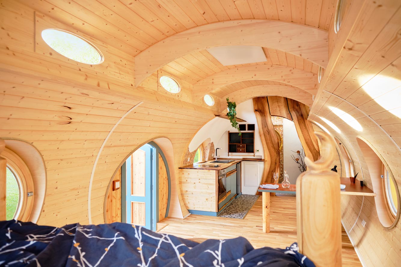 Innenraum eines handgefertigten Holzwagens mit gewölbter Holzdecke, runden Fenstern und kleiner Küchenzeile – Blick vom Bett Richtung Eingang